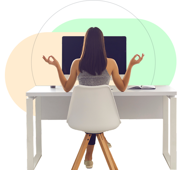 A woman is sitting with her back to us at a desktop computer. She is holding her hands in gyan mudra position, meditating.