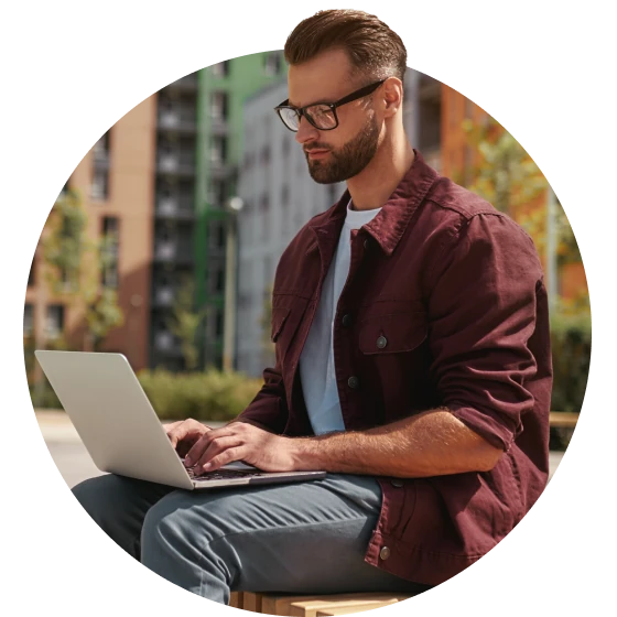 A man is sitting on a bench outside, browsing Usenet on his laptop. He is wearing a maroon button down shirt over a white t-shirt.